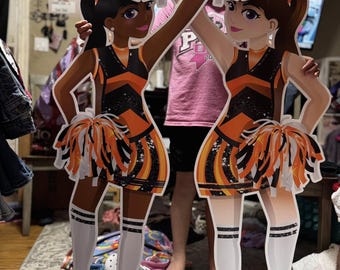 May include: Two cardboard cutouts of cheerleaders in orange, black, and white uniforms, holding orange and white pom-poms. The cheerleaders have dark hair and are wearing orange sneakers and knee-high socks. The cutouts are standing in front of a messy background.