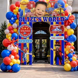 May include: A carnival-themed party entrance archway featuring a large balloon display in red, blue, and yellow. The structure includes banners reading BLAZE'S WORLD, WELCOME, and PRIZES, with illustrations of a clown and a ringmaster on the side panels.