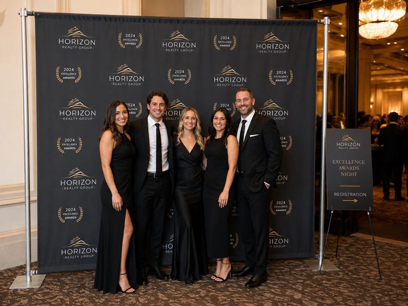 May include: A black step and repeat backdrop featuring the gold Horizon Realty Group logo and 2024 Excellence Awards text in a repeating pattern. A separate black sign on a stand displays Horizon Realty Group Excellence Awards Night Registration.