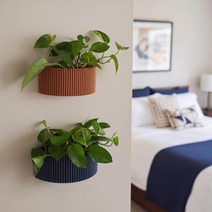 May include: Two semi-oval wall-mounted planters with a vertical ribbed texture. The top planter is terracotta orange and the bottom planter is navy blue. Each planter holds a green pothos plant with trailing heart-shaped leaves.