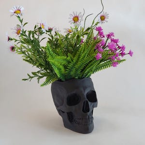May include: A black skull-shaped planter with a variety of white and pink flowers growing out of the top.