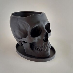 May include: A black skull-shaped planter with a round base. The planter is made of a smooth, matte material and has a detailed skull design.