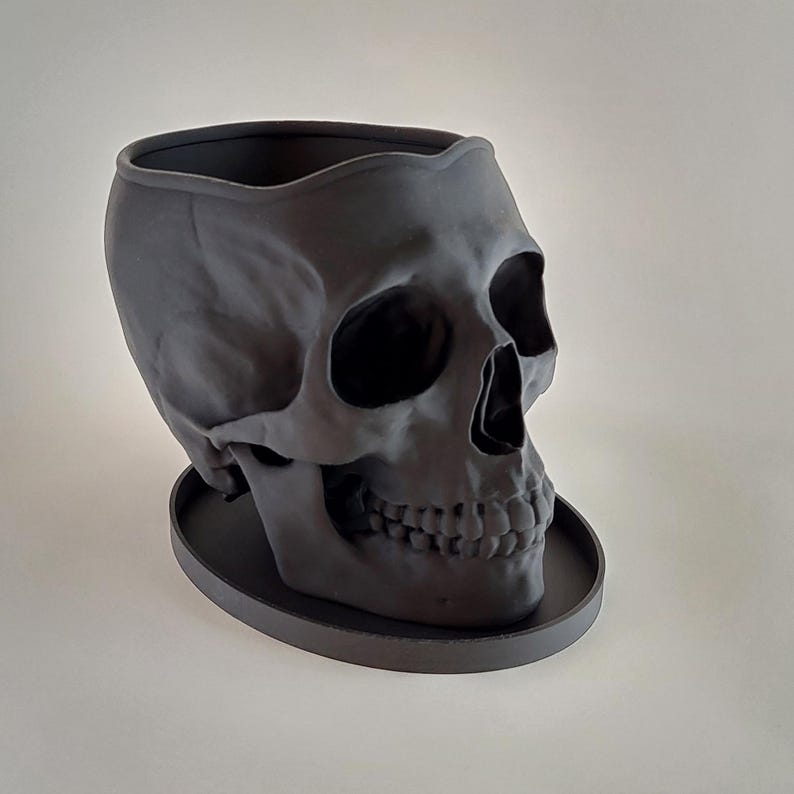 May include: A black skull-shaped planter with a round base. The planter is made of a smooth, matte material and has a detailed skull design.