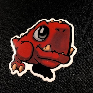 May include: A red cartoon dinosaur sticker with a white border. The dinosaur has a friendly expression with a large eye, tan teeth, and a smiling mouth. The sticker is on a black surface.