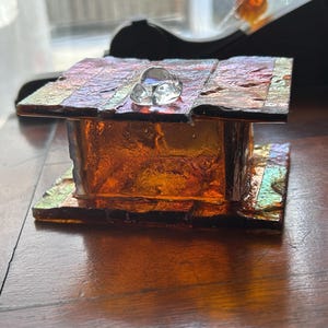 May include: A handcrafted, iridescent glass box with a clear glass accent on top. The box features a rectangular shape with a warm, amber color and a textured surface. The glass reflects multiple colors, creating a unique visual effect.