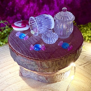 Könnte beinhalten: Zwei Miniatur-Gläser aus klarem Glas mit gerippter Textur und passenden Deckeln, eines aufrecht stehend und eines auf der Seite liegend, begleitet von mehreren kleinen, bonbonförmigen Anhängern in Blau und Pink auf einer runden Holzscheibe.