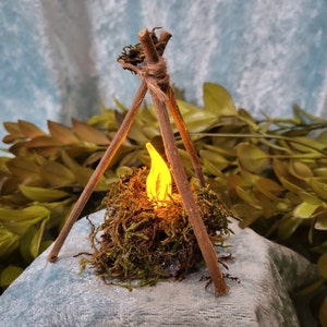 Könnte beinhalten: Eine Miniatur-Lagerfeuerszene mit einer künstlichen Flamme. Das Feuer besteht aus einem Bündel grünen Mooses und Zweigen, mit einem kleinen, gelben LED-Licht, das eine Flamme simuliert. Das Feuer wird von drei Holzstäben getragen, die oben zusammengebunden sind.