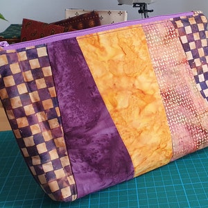 Könnte beinhalten: Eine gepolsterte, mit einem Reißverschluss versehene Tasche in Lila und Braun mit einem karierten Muster auf einer Seite. Die Tasche ist aus Stoff mit einem Batik-Muster.