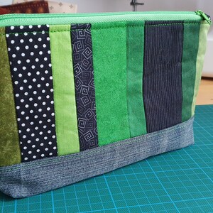 Könnte beinhalten: Eine grüne und schwarze gestreifte Reißverschlusstasche mit einem Jeansboden. Die Tasche besteht aus verschiedenen Stoffen mit verschiedenen Mustern, darunter Punkte, geometrische Formen und Streifen.