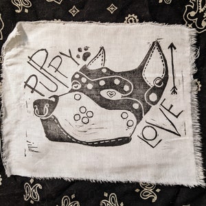 May include: A white fabric square with a black linocut print of a dog's face, featuring the words "PUPPY LOVE" and an arrow. The dog's face has dotted patterns and a heart.