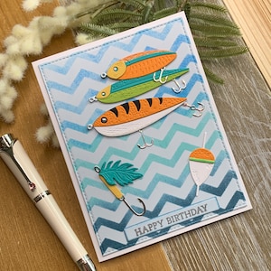 May include: A handmade birthday card featuring colorful fishing lures. The card has a light blue chevron pattern background with the words "Happy Birthday" at the bottom. The lures are orange, green, and striped, with silver hooks.