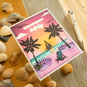 May include: A birthday card with a beach theme, featuring a sunset, palm trees, a sailboat, and a beach chair. The card includes the text "Relax and enjoy your birthday". Seashells and a pen are visible in the background, creating a coastal atmosphere.