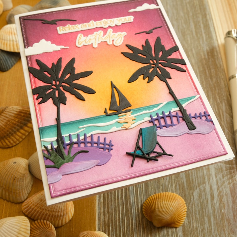 May include: A handmade birthday card depicting a beach scene with palm trees, a sailboat, and a deckchair. The card has a sunset background with the words "Relax and enjoy your birthday". Seashells surround the card.