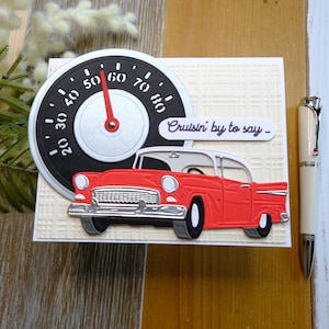 May include: A greeting card featuring a red classic car with a white roof, a speedometer, and the phrase "Cruisin' by to say -". The card has a textured background and a pen is placed on the right side.