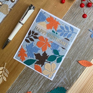 May include: A handmade birthday card with embossed leaves in shades of orange, blue, brown, and green. The card has the words "HAPPY BIRTHDAY!" in gold lettering. A white pen with silver accents rests on the wooden surface.