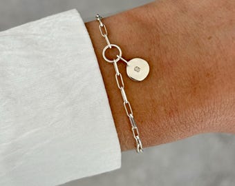 May include: A silver bracelet with a delicate chain design. The bracelet features a small, round charm with a single, embedded diamond. The charm is connected to the chain by a small ring. The bracelet is worn on a wrist.