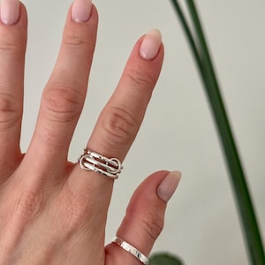 May include: Two silver rings are shown on a hand. One ring features a distinctive, layered design, and the other is a simple band with a textured surface. The rings are set against a neutral background with a touch of greenery.