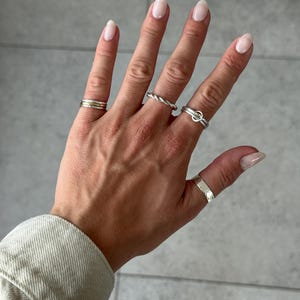 May include: A collection of four silver-toned rings worn on the fingers. One ring features a multi-band design, another has a twisted rope texture, one includes a small circular charm, and the thumb ring has a wide, hammered, textured surface.