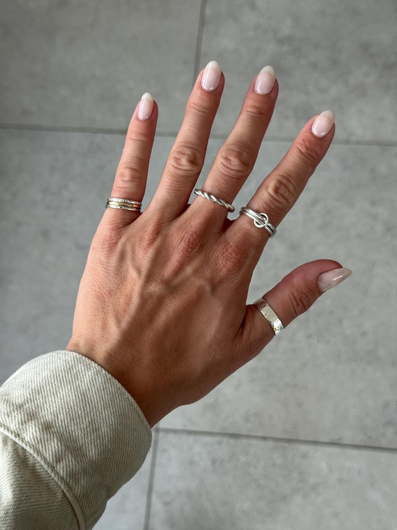 May include: A collection of four silver-toned rings worn on the fingers. One ring features a multi-band design, another has a twisted rope texture, one includes a small circular charm, and the thumb ring has a wide, hammered, textured surface.
