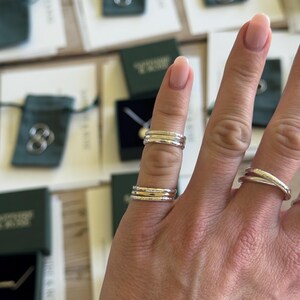 May include: A set of stacked rings featuring a combination of silver and gold-toned bands. The middle finger displays two separate stacks of three thin rings each, while the ring finger features a single crossover band with a polished finish.