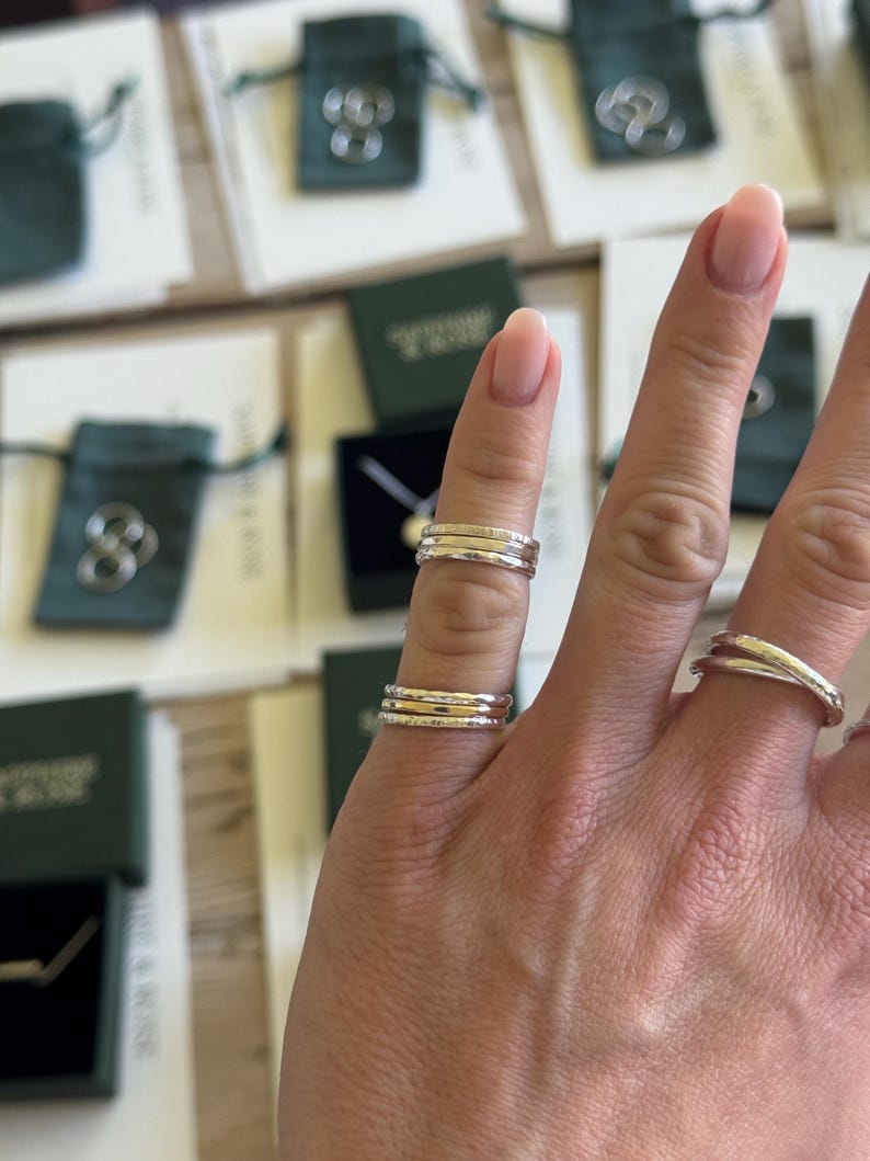 May include: A set of stacked rings featuring a combination of silver and gold-toned bands. The middle finger displays two separate stacks of three thin rings each, while the ring finger features a single crossover band with a polished finish.