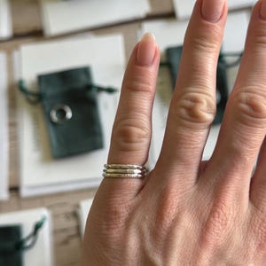 May include: A set of three thin, stacked silver rings worn on a finger. Each ring features a distinct texture, with one smooth band, one hammered band, and one band with a subtle, irregular finish, all crafted from polished silver metal.
