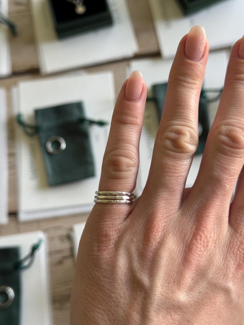 May include: A set of three thin, stacked silver rings worn on a finger. Each ring features a distinct texture, with one smooth band, one hammered band, and one band with a subtle, irregular finish, all crafted from polished silver metal.