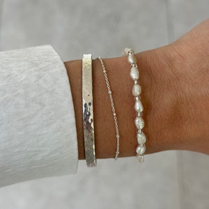 May include: Three silver bracelets on a wrist. The bracelet on the bottom is a wide, hammered band. The middle bracelet is a delicate silver chain. The top bracelet is made of white pearls.