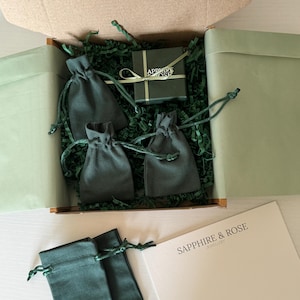 May include: A cardboard shipping box containing green shredded paper, three dark green drawstring pouches, and a small dark green gift box tied with a light green ribbon. Outside the box are two additional green pouches and a white card with the text SAPPHIRE & ROSE JEWELLERY.