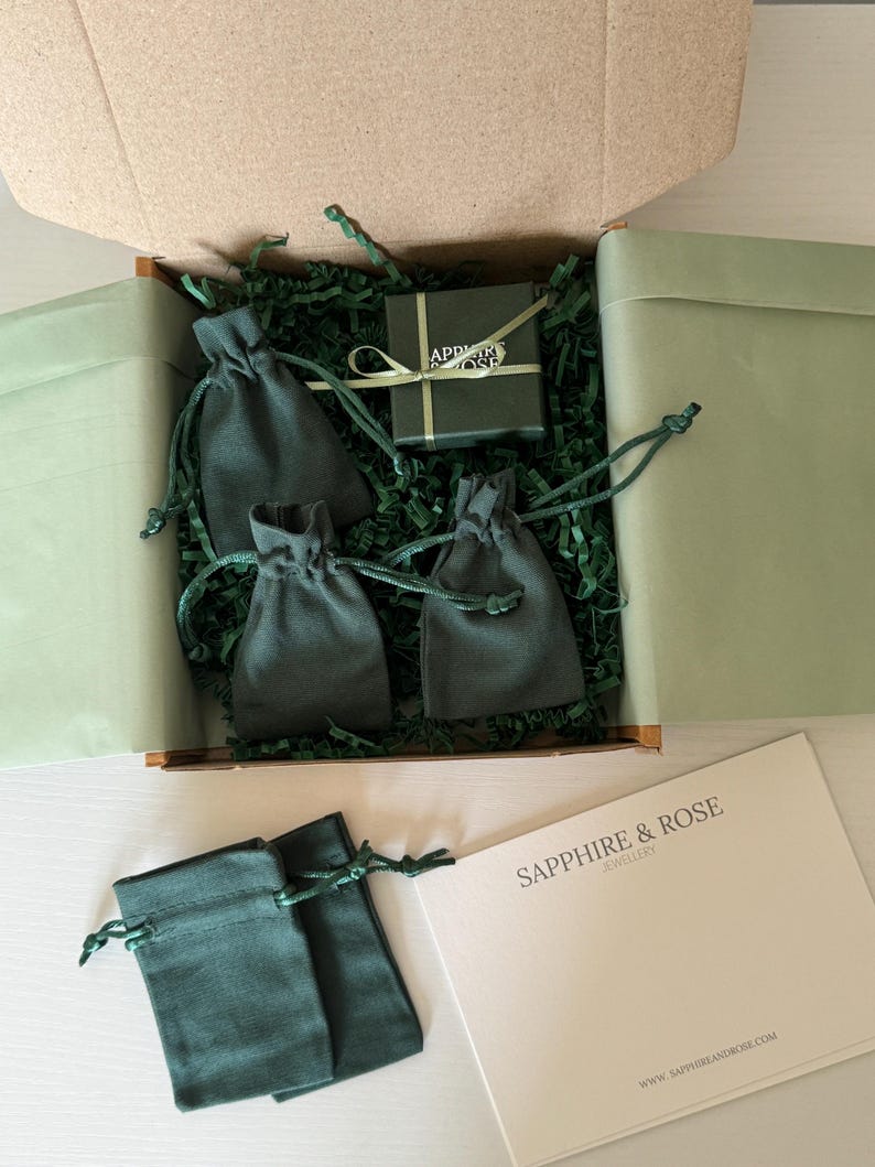 May include: A cardboard shipping box containing green shredded paper, three dark green drawstring pouches, and a small dark green gift box tied with a light green ribbon. Outside the box are two additional green pouches and a white card with the text SAPPHIRE & ROSE JEWELLERY.