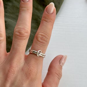 May include: A silver ring with two bands, one of which is twisted into a knot. The ring is worn on a person's finger.