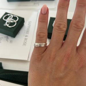 May include: A set of three thin, silver-coloured stacking rings worn on a finger. Each ring features a distinct texture, with one smooth band, one hammered band, and one band with a subtle, repeating pattern, all crafted from a metallic material.