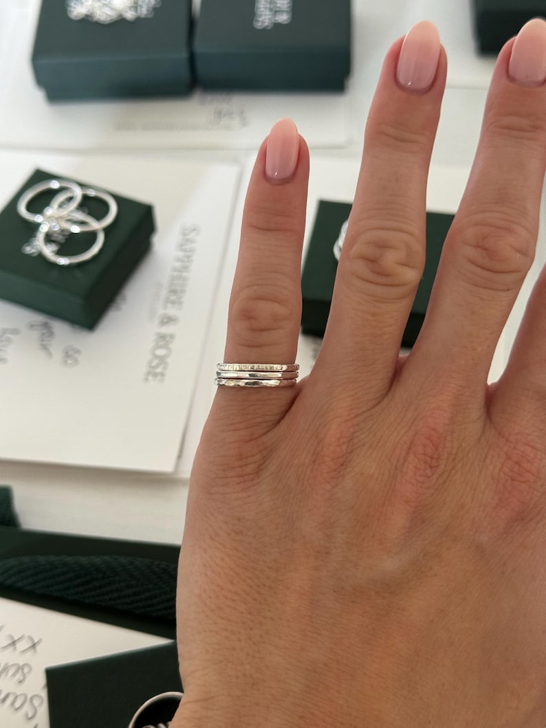 May include: A set of three thin, silver-coloured stacking rings worn on a finger. Each ring features a distinct texture, with one smooth band, one hammered band, and one band with a subtle, repeating pattern, all crafted from a metallic material.