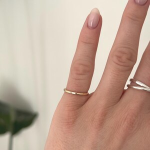 Pode incluir: Um anel de banda de ouro polido fino usado no dedo m&eacute;dio, apresentando uma superf&iacute;cie lisa e arredondada com um acabamento met&aacute;lico quente. O anel tem um design simples, exibindo uma forma circular consistente que se ajusta ao dedo.