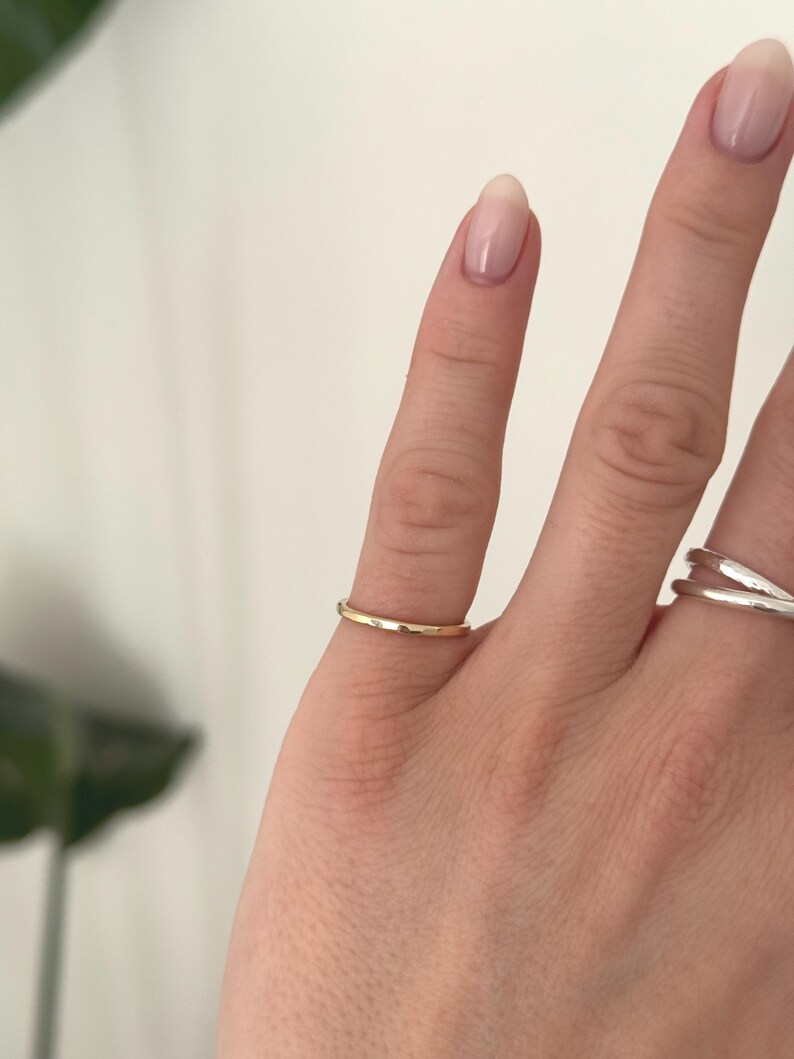 Pode incluir: Um anel de banda de ouro polido fino usado no dedo m&eacute;dio, apresentando uma superf&iacute;cie lisa e arredondada com um acabamento met&aacute;lico quente. O anel tem um design simples, exibindo uma forma circular consistente que se ajusta ao dedo.