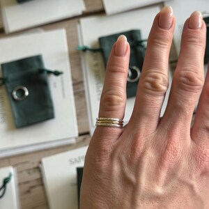 Pode incluir: Um conjunto de tr&ecirc;s an&eacute;is finos empilhados num dedo. O conjunto consiste em dois an&eacute;is exteriores com um acabamento prateado martelado e texturizado, e um anel central com um acabamento dourado liso e polido, usados em conjunto.