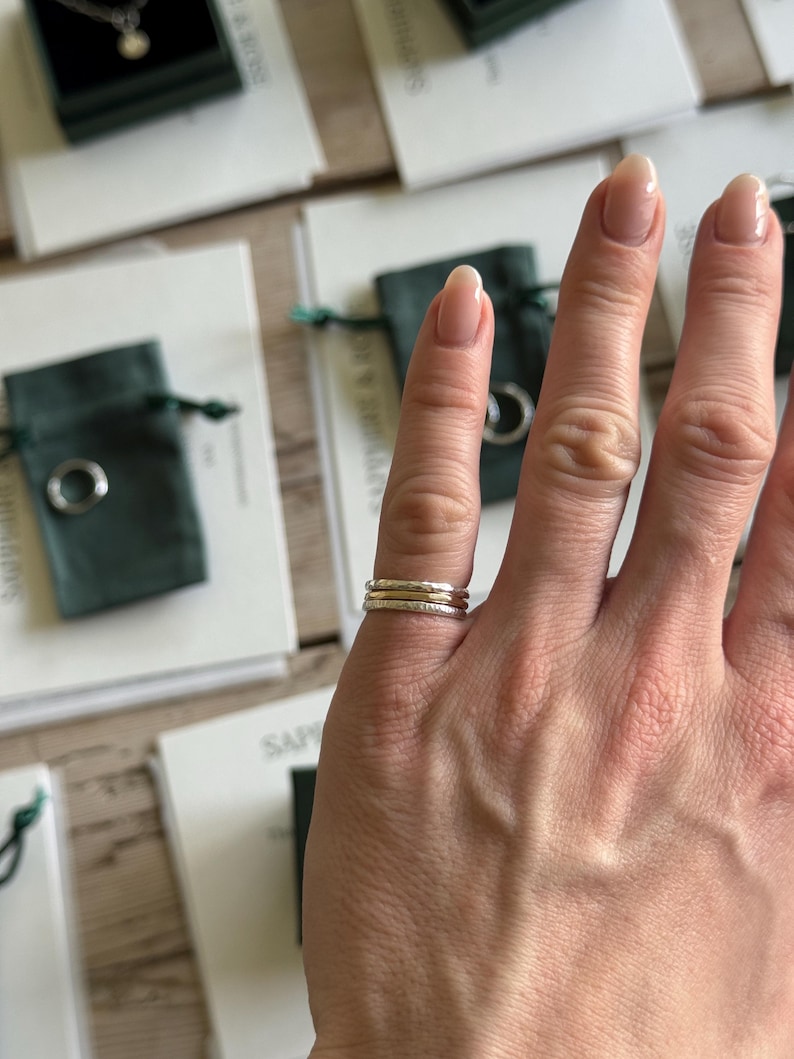 Pode incluir: Um conjunto de tr&ecirc;s an&eacute;is finos empilhados num dedo. O conjunto consiste em dois an&eacute;is exteriores com um acabamento prateado martelado e texturizado, e um anel central com um acabamento dourado liso e polido, usados em conjunto.