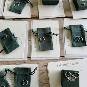 May include: A collection of silver jewellery pieces including rings and hoop earrings displayed with dark green fabric drawstring pouches and dark green gift boxes, all accompanied by white cards printed with the text Sapphire & Rose Jewellery.