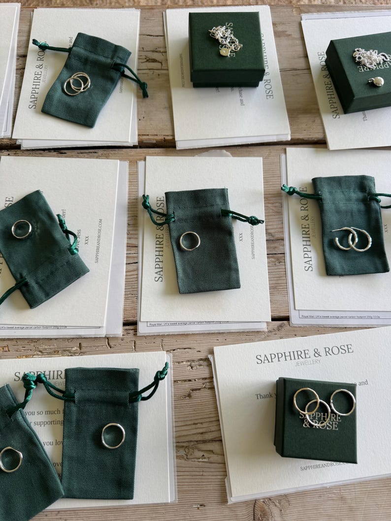 May include: A collection of silver jewellery pieces including rings and hoop earrings displayed with dark green fabric drawstring pouches and dark green gift boxes, all accompanied by white cards printed with the text Sapphire & Rose Jewellery.