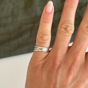 May include: A set of three stacked silver rings worn on a finger. The rings feature different textures, including one smooth band and two bands with a hammered, irregular surface finish that reflects light across the metallic silver material.