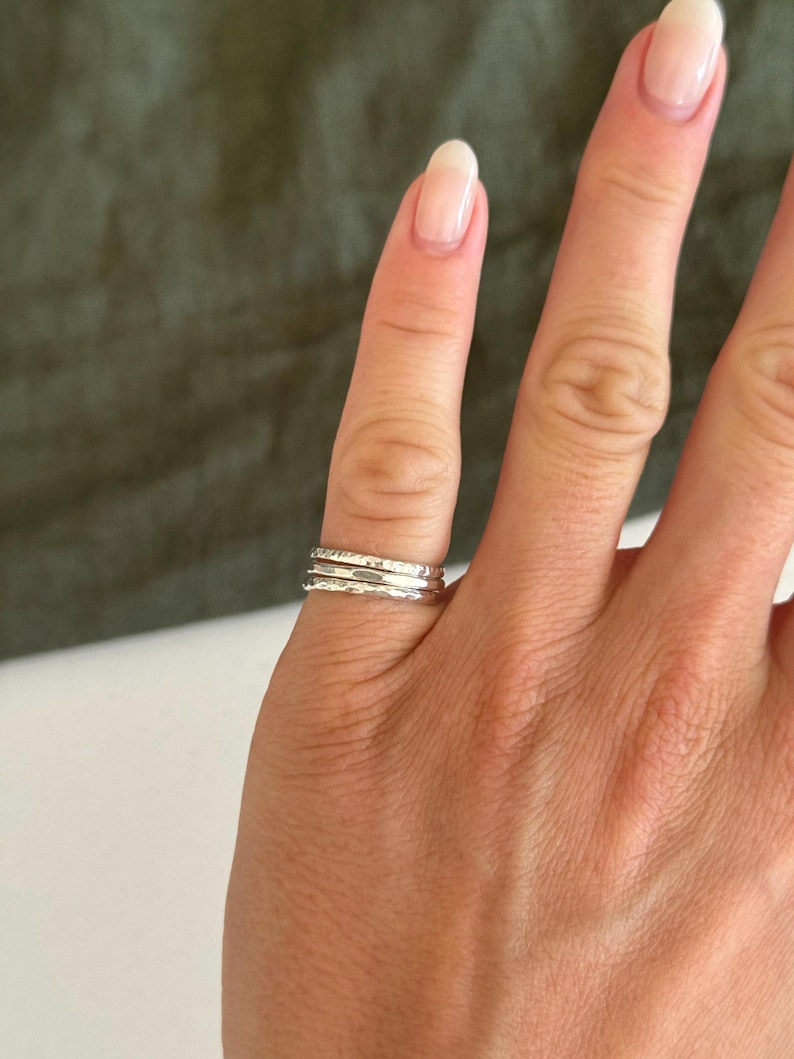 May include: A set of three stacked silver rings worn on a finger. The rings feature different textures, including one smooth band and two bands with a hammered, irregular surface finish that reflects light across the metallic silver material.