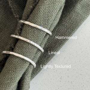 May include: Three silver-toned rings with distinct surface finishes. The top ring is labelled Hammered, the middle ring is labelled Lineal, and the bottom ring is labelled Lightly Textured. The rings are arranged vertically on a folded olive green fabric.