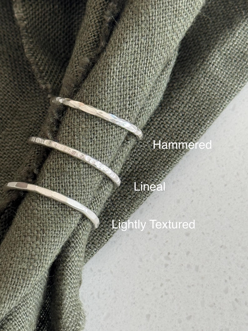 May include: Three silver-toned rings with distinct surface finishes. The top ring is labelled Hammered, the middle ring is labelled Lineal, and the bottom ring is labelled Lightly Textured. The rings are arranged vertically on a folded olive green fabric.