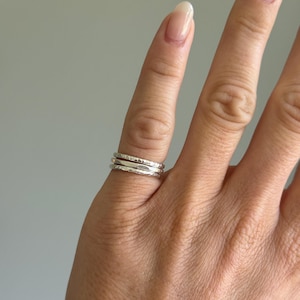 May include: A set of three thin, stacked rings worn on a finger. Each ring features a distinct metallic finish, with two bands displaying a textured, hammered surface and one band featuring a smooth, polished silver appearance.