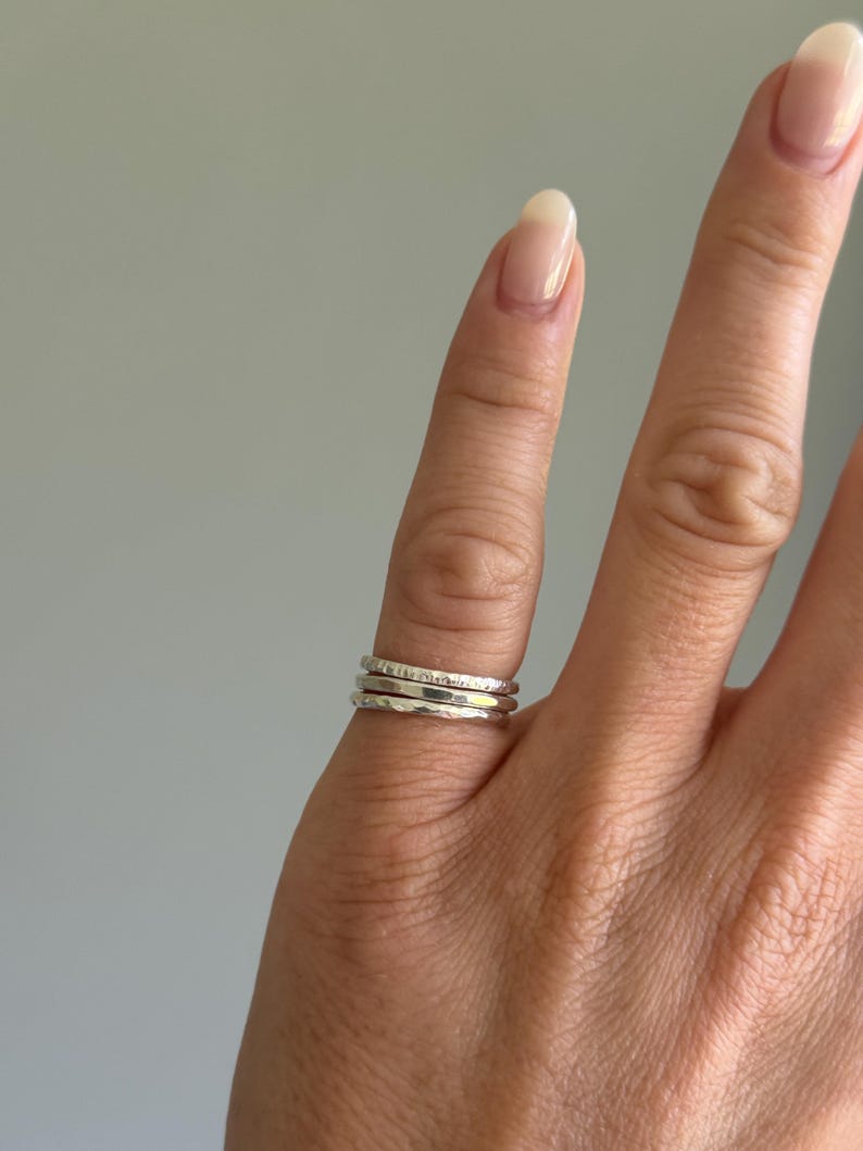 May include: A set of three thin silver-coloured stacking rings worn on a finger. The rings feature different textures, including one with a hammered finish, one with a smooth polished surface, and one with a subtle, fine-grained or etched pattern.