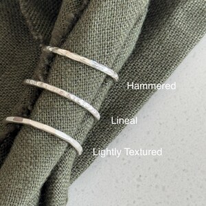 Pode incluir: Tr&ecirc;s an&eacute;is finos de prata com acabamentos de superf&iacute;cie distintos. O anel superior est&aacute; rotulado como Hammered, o anel do meio como Lineal e o anel inferior como Lightly Textured. Os an&eacute;is est&atilde;o dispostos verticalmente sobre um tecido verde-oliva.