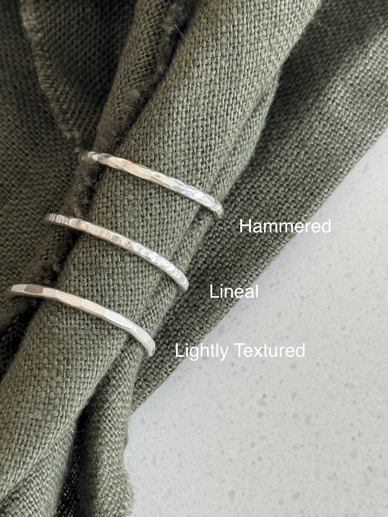 Pode incluir: Tr&ecirc;s an&eacute;is finos de prata com acabamentos de superf&iacute;cie distintos. O anel superior est&aacute; rotulado como Hammered, o anel do meio como Lineal e o anel inferior como Lightly Textured. Os an&eacute;is est&atilde;o dispostos verticalmente sobre um tecido verde-oliva.