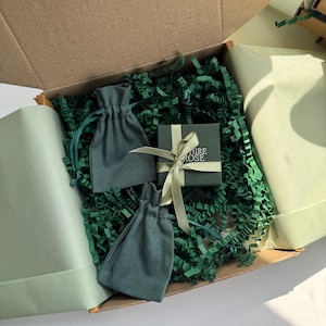 Pode incluir: Uma caixa de envio de papel&atilde;o contendo duas bolsas de tecido verde-s&aacute;lvia com cord&atilde;o e uma pequena caixa de presente quadrada verde-escura amarrada com uma fita verde-clara. A caixa tem o texto "SAPPHIRE ROSE" impresso em branco na tampa.