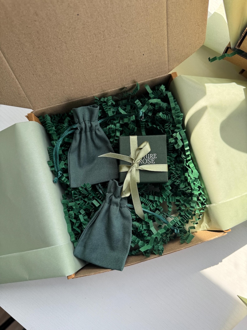Pode incluir: Uma caixa de envio de papel&atilde;o contendo duas bolsas de tecido verde-s&aacute;lvia com cord&atilde;o e uma pequena caixa de presente quadrada verde-escura amarrada com uma fita verde-clara. A caixa tem o texto "SAPPHIRE ROSE" impresso em branco na tampa.