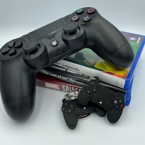 Könnte beinhalten: Ein schwarzer PlayStation 4-Controller mit einer schwarzen PlayStation 4-Konsole und drei übereinander gestapelten Videospiel-Hüllen. Die Videospiel-Hüllen sind für "The Last of Us Remastered", "Marvel's Spider-Man" und "God of War".